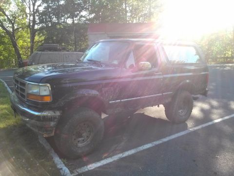 1995 Ford Bronco - This muddy Ford Bronco is parked in a lot, with trees and a building in the background.