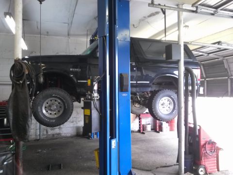 1995 Ford Bronco - A black 1995 Ford Bronco is raised on a hydraulic lift inside a garage, revealing its undercarriage and off-road tires.