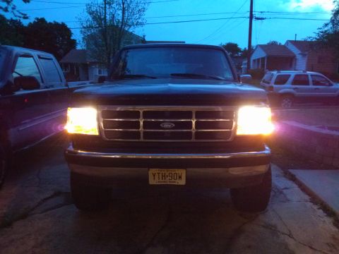 1995 Ford Bronco - The front grille and headlights of a 1995 Ford Bronco are visible, with another vehicle to the left and an SUV in the background.