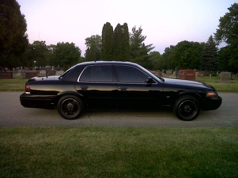 1999 Ford Crown Victoria - Side view of a black sedan parked on a road, with trees and gravestones visible.
