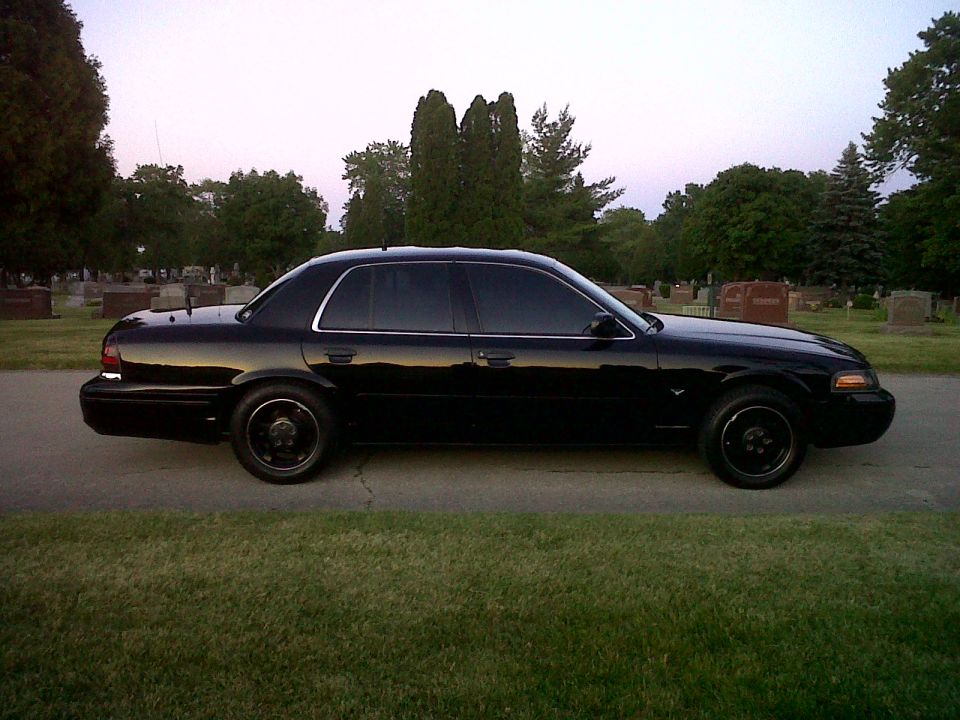 Black sedan parked on a road with trees and gravestones in the background