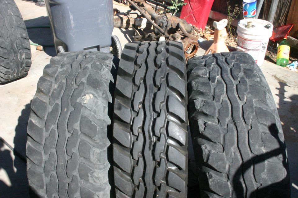 Three off-road tires lined up, with a gray trash can and vehicle parts in the background