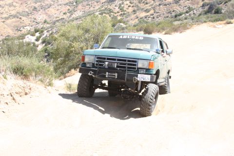 1991 Ford Bronco - A teal Ford Bronco with off-road modifications drives on a dirt path in a dry, hilly landscape.