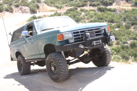 1991 Ford Bronco - 1991 Ford Bronco navigates an off-road trail with large tires and modified front end.