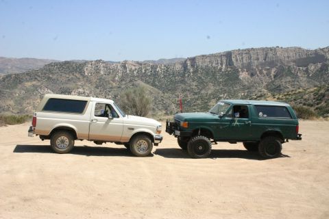 1991 Ford Bronco - Two Ford Broncos, a white one and a green one, are parked side-by-side in a dry, mountainous landscape.