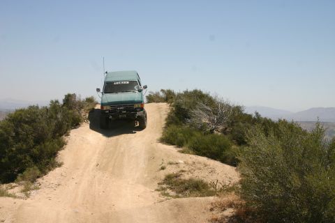 1991 Ford Bronco - A teal SUV navigates a dusty off-road trail under a clear blue sky.