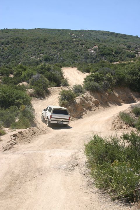 1991 Ford Bronco - A white SUV navigates a dusty, off-road trail through a landscape of green hills and sparse vegetation.