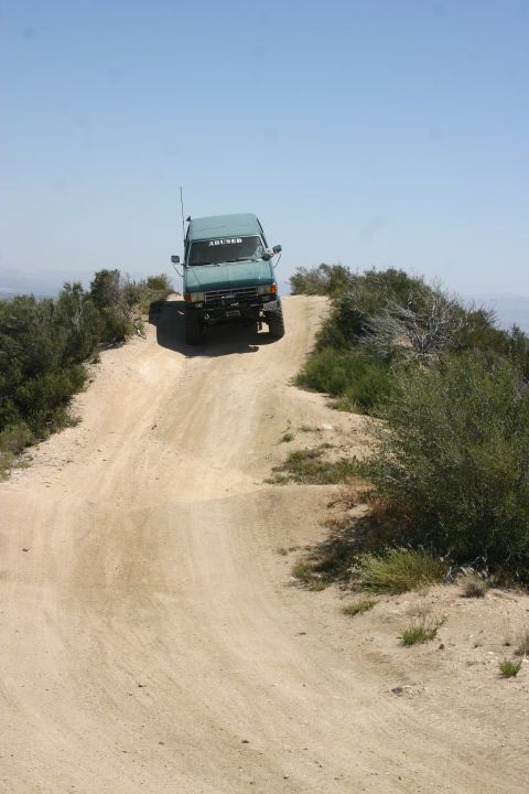 1991 Ford Bronco - This teal SUV, with "ABUSED" emblazoned on its windshield, navigates a steep dirt incline on a sunny day.