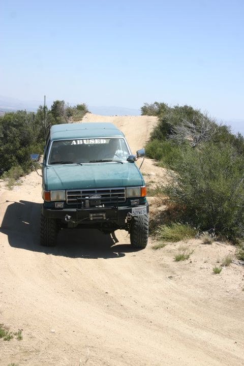 1991 Ford Bronco - Teal SUV with winch and off-road tires navigates a dusty trail, sporting an "ABUSED" sticker on the windshield.