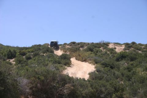 1991 Ford Bronco - A dark SUV navigates a steep dirt path through a scrub-covered hillside on a bright, clear day.