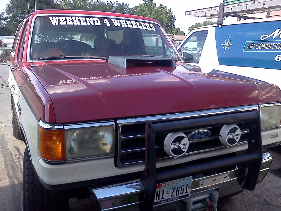 1989 Ford Bronco front view with aftermarket hood scoop and "WEEKEND 4 WHEELERS" windshield banner. Another vehicle is partially visible to the right
