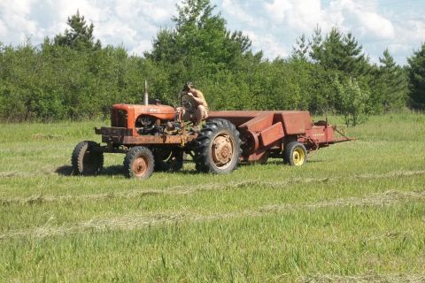 1987 Ford F-250 - A person operates an orange tractor attached to a red baler, working in a field of cut grass.