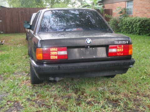 1991 BMW 3 Series - A black BMW 325i sedan is seen from the rear, parked outdoors on a grassy area with another vehicle partially visible behind it.