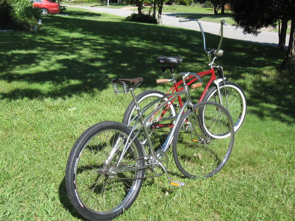 Two cruiser bicycles on grass, one chrome and one red, with a red car in the background