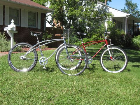 1994 Ford Bronco - Two classic cruiser bicycles, one in chrome and one in red, are parked on a sunny lawn in front of suburban houses.