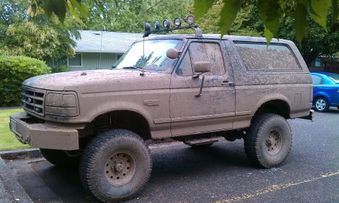 1995 Ford Bronco - This heavily mud-splattered Ford Bronco is parked on asphalt, with a blue car visible in the background.