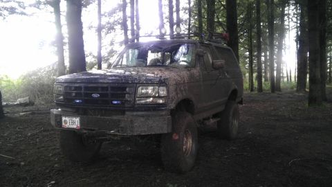 1995 Ford Bronco - This SUV is covered in mud and parked in a wooded area with sunlight filtering through the trees.