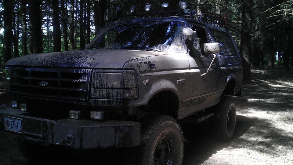 Mud-covered 1992 Ford Bronco with off-road lights on roof rack, parked in a forest