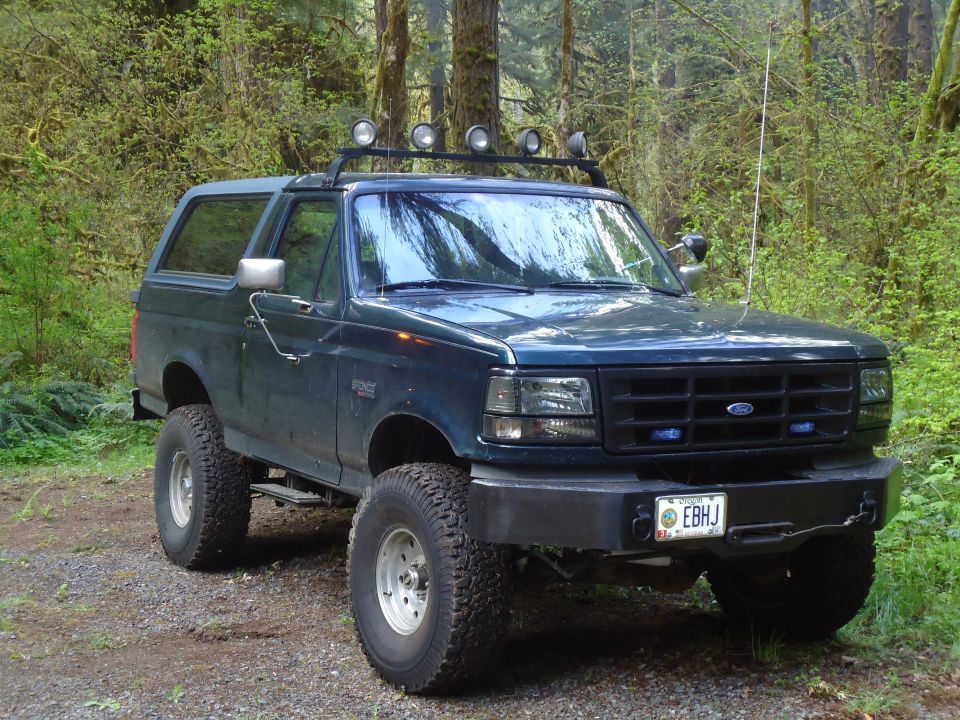 Dark blue Ford Bronco with roof lights and off-road tires parked on a dirt road in a forest