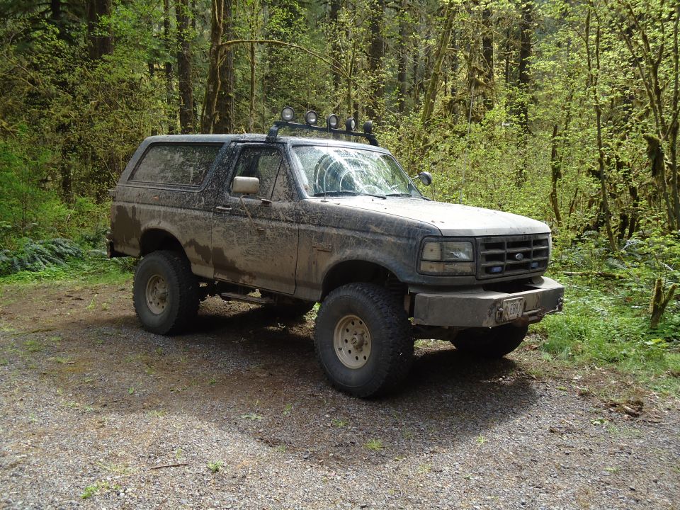 Mud-covered dark SUV with off-road tires and roof lights parked on a gravel road in a forest
