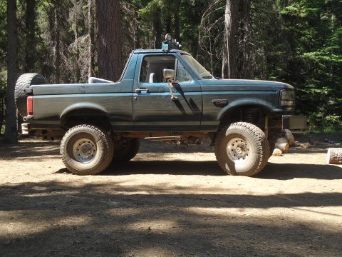 1995 Ford Bronco - This lifted 1995 Ford Bronco is parked in a wooded area, showing its side profile and spare tire mounted on the rear.