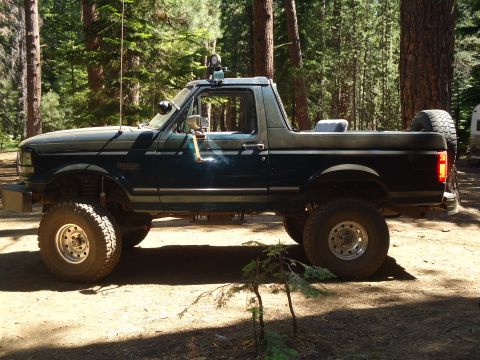 1995 Ford Bronco - A lifted 1995 Ford Bronco is parked in a wooded area, showing its side profile and off-road modifications.