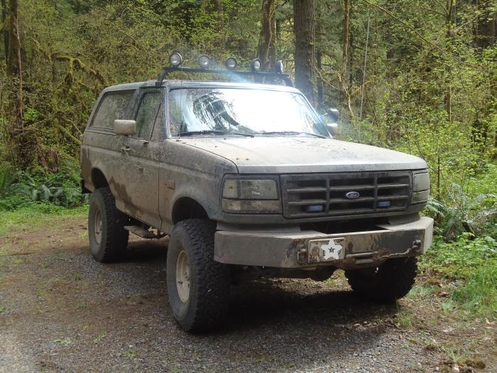 Muddy Ford Bronco parked on a dirt road in a forest. It has a light bar on the roof and a custom bumper