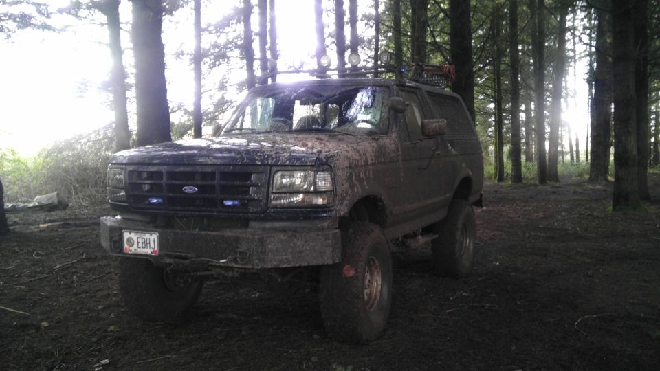 1995 Ford Bronco covered in mud, parked in a forest with trees and sunlight