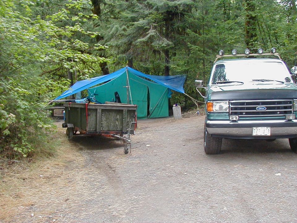 Green Ford van, front view, parked at campsite with trailer and tent. License plate visible