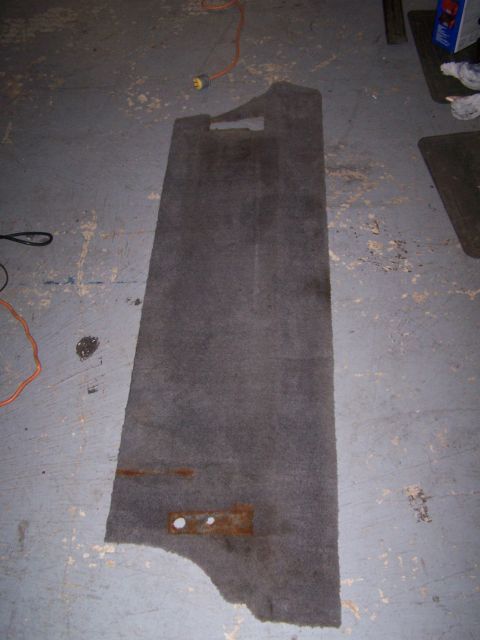 1995 Ford Bronco - A gray carpeted floor mat with visible rust stains and mounting holes lies on a concrete floor.