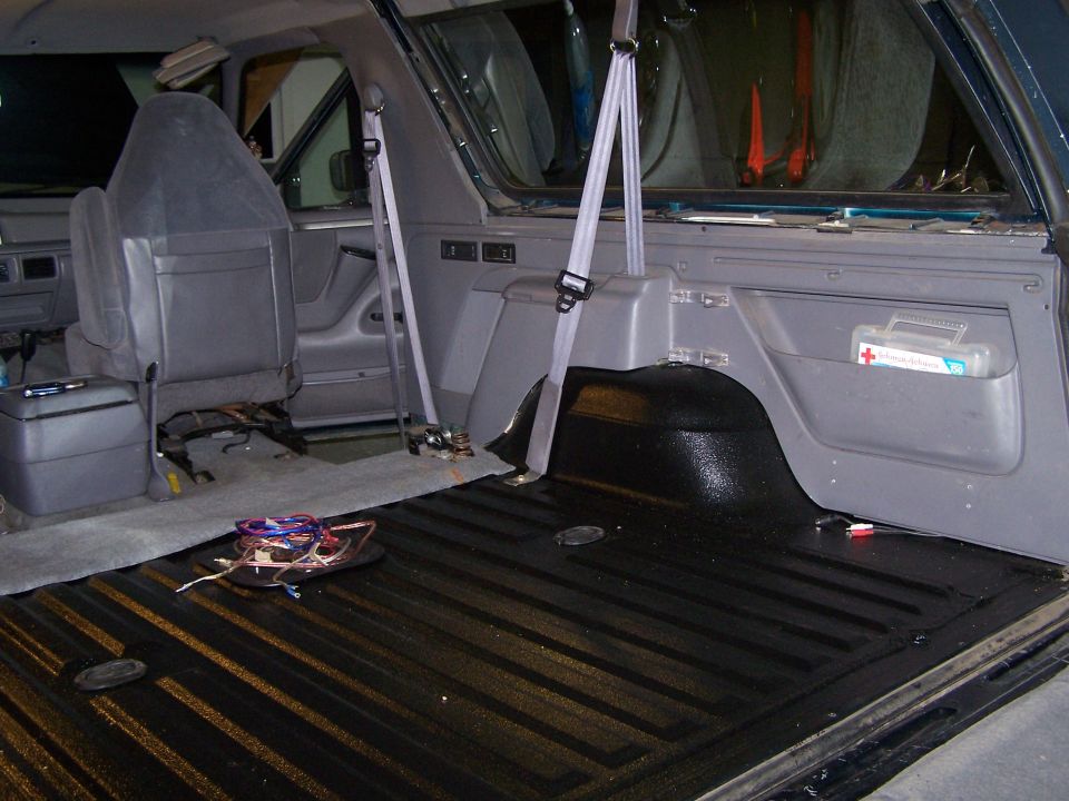 Interior view of a vehicle's cargo area with seats folded down, showing a black ribbed floor, seatbelts, and a first-aid kit in a side pocket