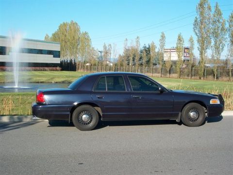 2001 Ford Crown Victoria - A dark blue sedan is parked on the side of the road with a fountain and building in the background.