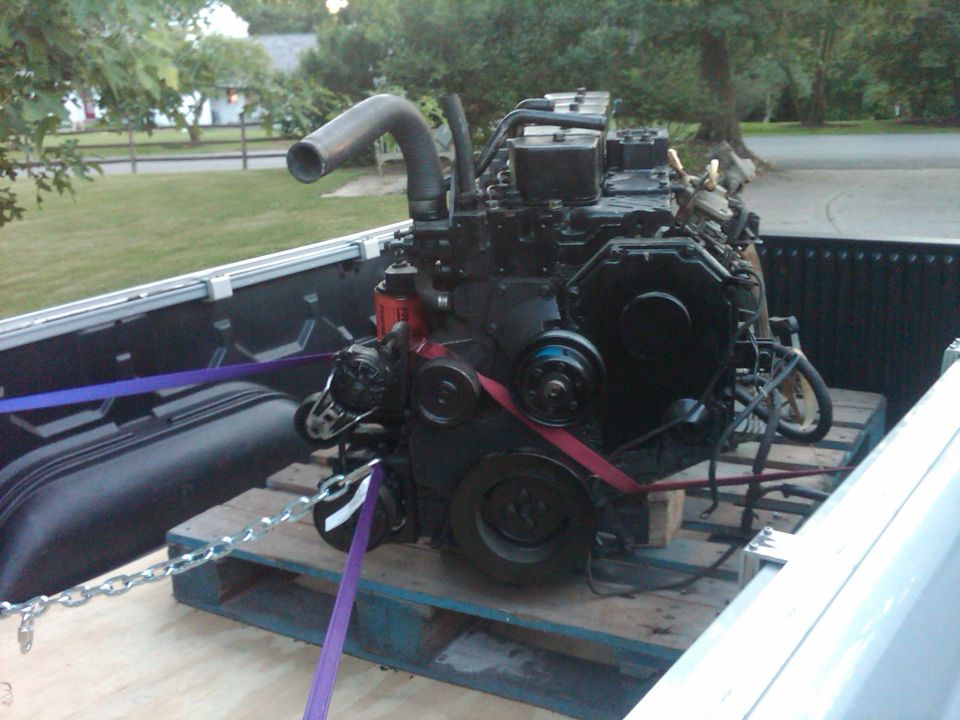 Black diesel engine on wooden pallets in truck bed, secured with purple straps and chains