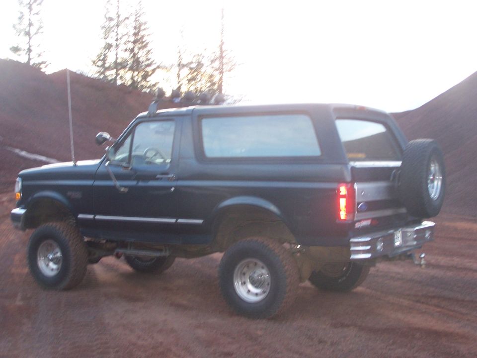 Dark blue Ford Bronco with a lift kit parked on a dirt road, spare tire mounted on the back
