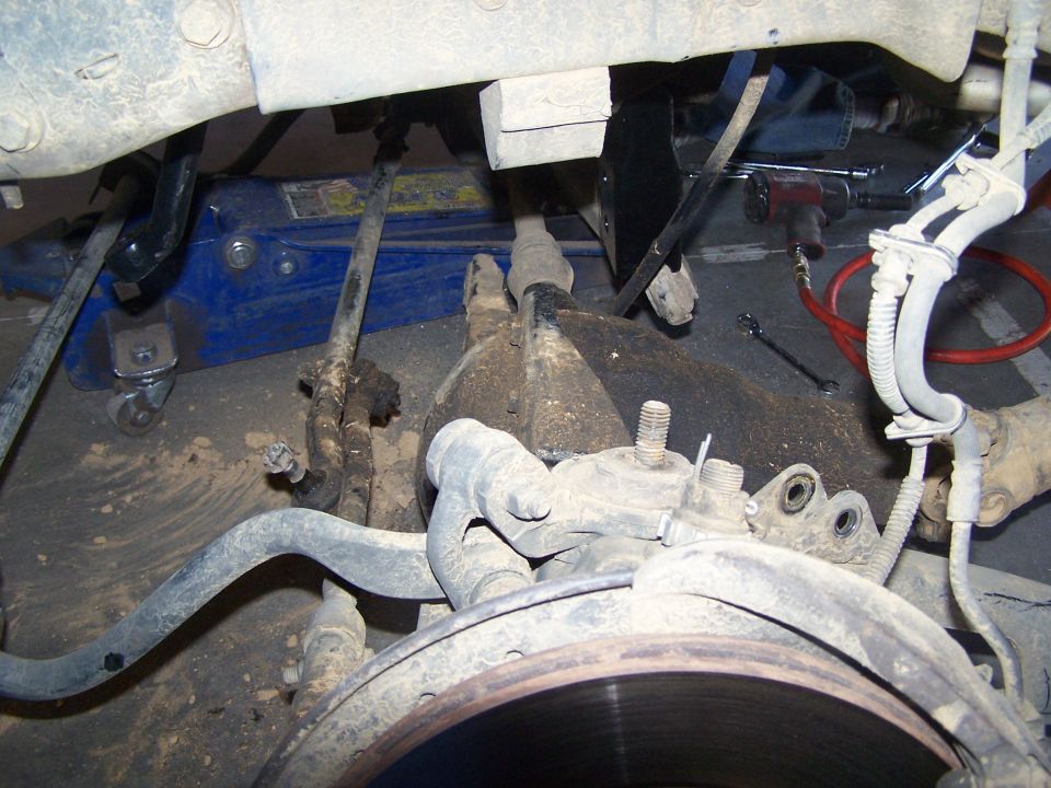 Vehicle suspension components with a blue hydraulic jack underneath. Visible parts include brake rotor, control arm, and steering linkage