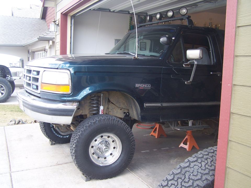 Dark green Ford Bronco with a lift kit, jacked up in a garage. It has off-road tires and a roof rack with lights