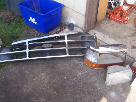 1995 Ford Bronco - Disassembled grille and headlight components for a 1992 Ford Bronco are laid out on the ground.
