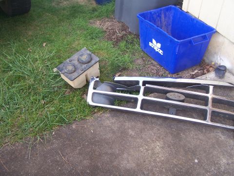 1995 Ford Bronco - A chrome grille and plastic fluid reservoir, likely from a 1995 Ford Bronco, lie on the grass next to a blue bin.
