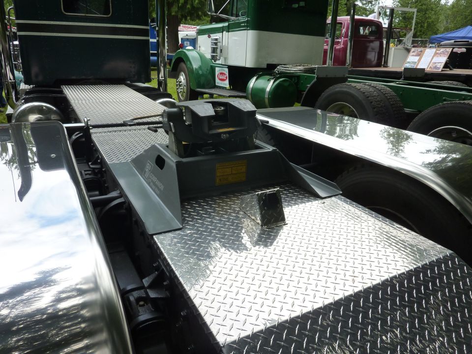 Close-up of a semi-truck's fifth wheel hitch and diamond plate deck. Another truck is visible behind it