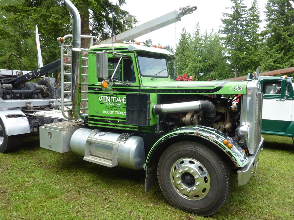 Green logging truck with crane, "VINTAGE TRUCKING" text on door. Another truck is partially visible to the right