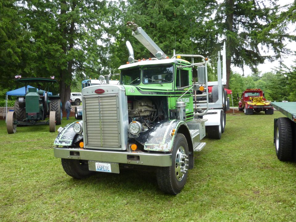 Green logging truck with crane, Peterbilt badge on grille. Other trucks and a tractor are visible in the background