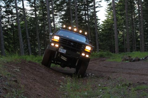 1995 Ford Bronco - This black Ford Bronco, likely a 1992 model, navigates a wooded trail with its auxiliary lights illuminated.