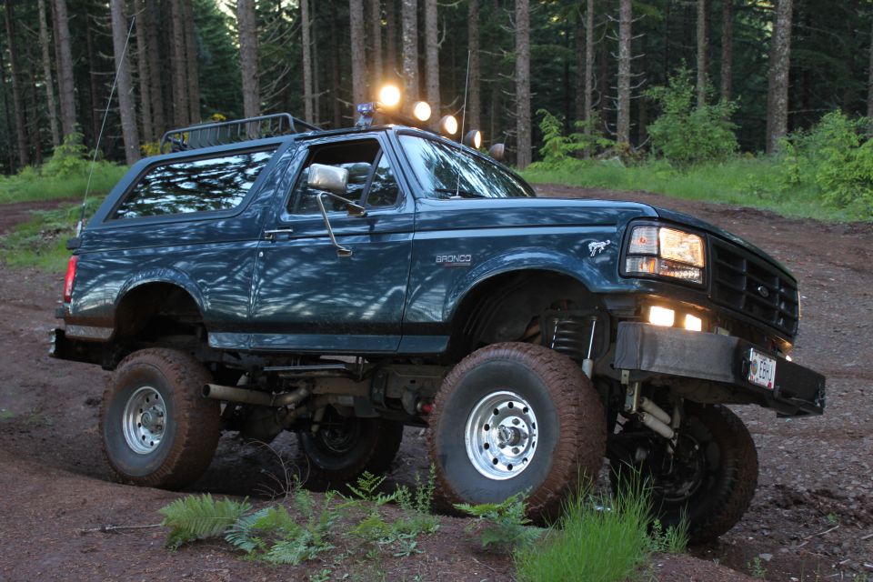 Dark teal lifted Ford Bronco with off-road tires on a dirt path in a forest