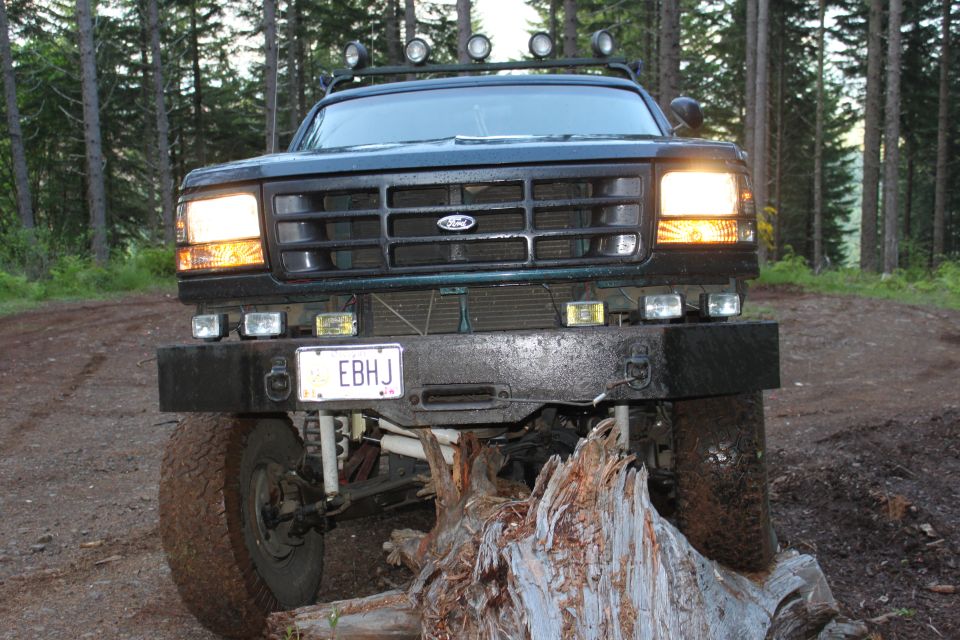 Dark green Ford Bronco with a winch and aftermarket bumper, front view, on a dirt road with trees