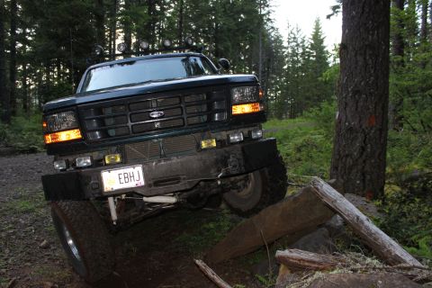 1995 Ford Bronco - A dark green Ford truck navigates a rocky, wooded trail with logs obstructing the path.