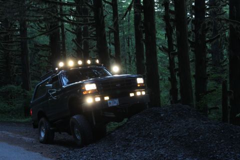 1995 Ford Bronco - A black 1995 Ford Bronco with multiple roof lights navigates a rocky incline in a dark forest setting.