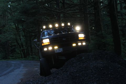 1995 Ford Bronco - Ford truck with multiple auxiliary lights on the roof and bumper, navigating a wooded area at dusk.