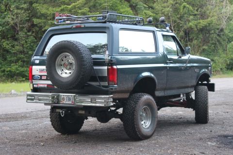 1995 Ford Bronco - This lifted Ford Bronco is parked on a dirt road with trees in the background.