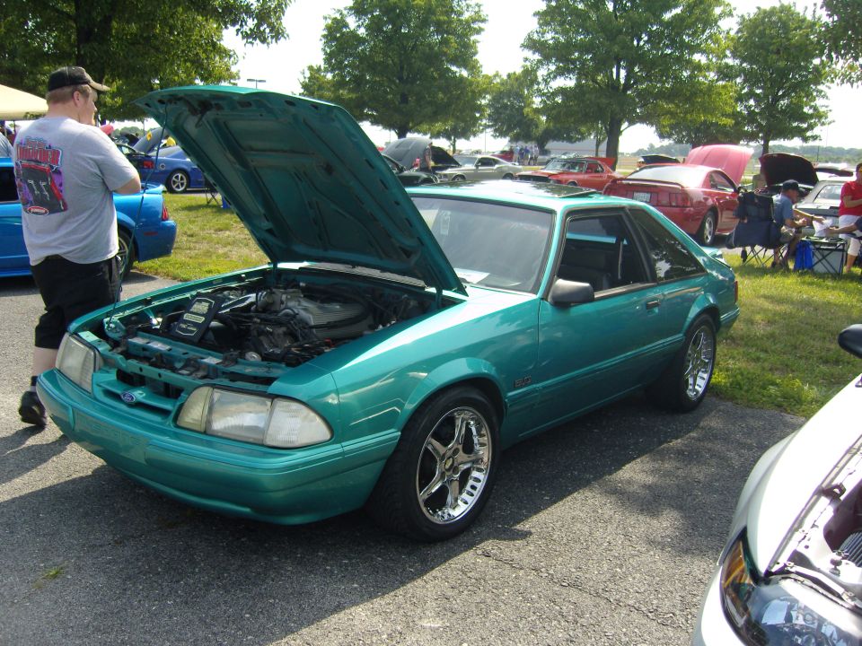 Teal 1990 Ford Mustang with hood open at a car show. Another car is partially visible in the foreground