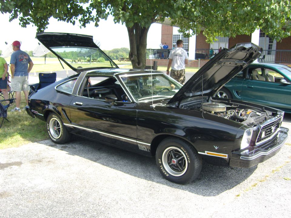 Black 1970s Ford Mustang fastback with hood and trunk open, parked outside. Another green car is visible to the right